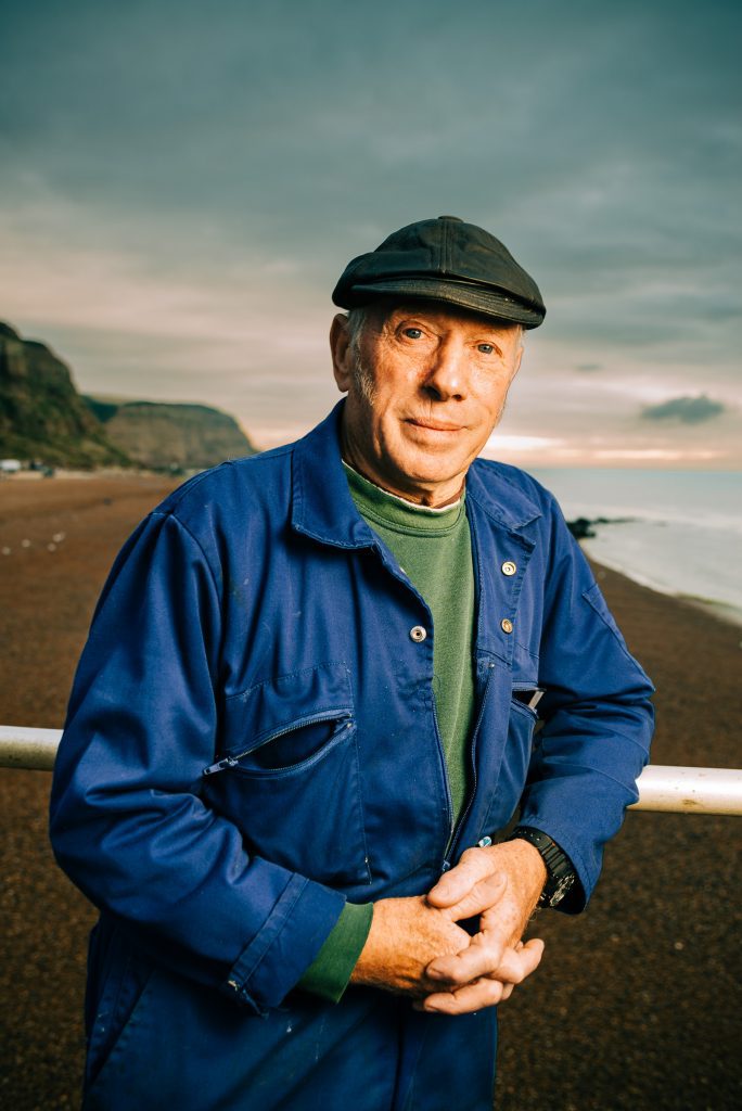 Environmental portrait of Hastings fisherman in blue work jacket and flat cap on Sussex coast